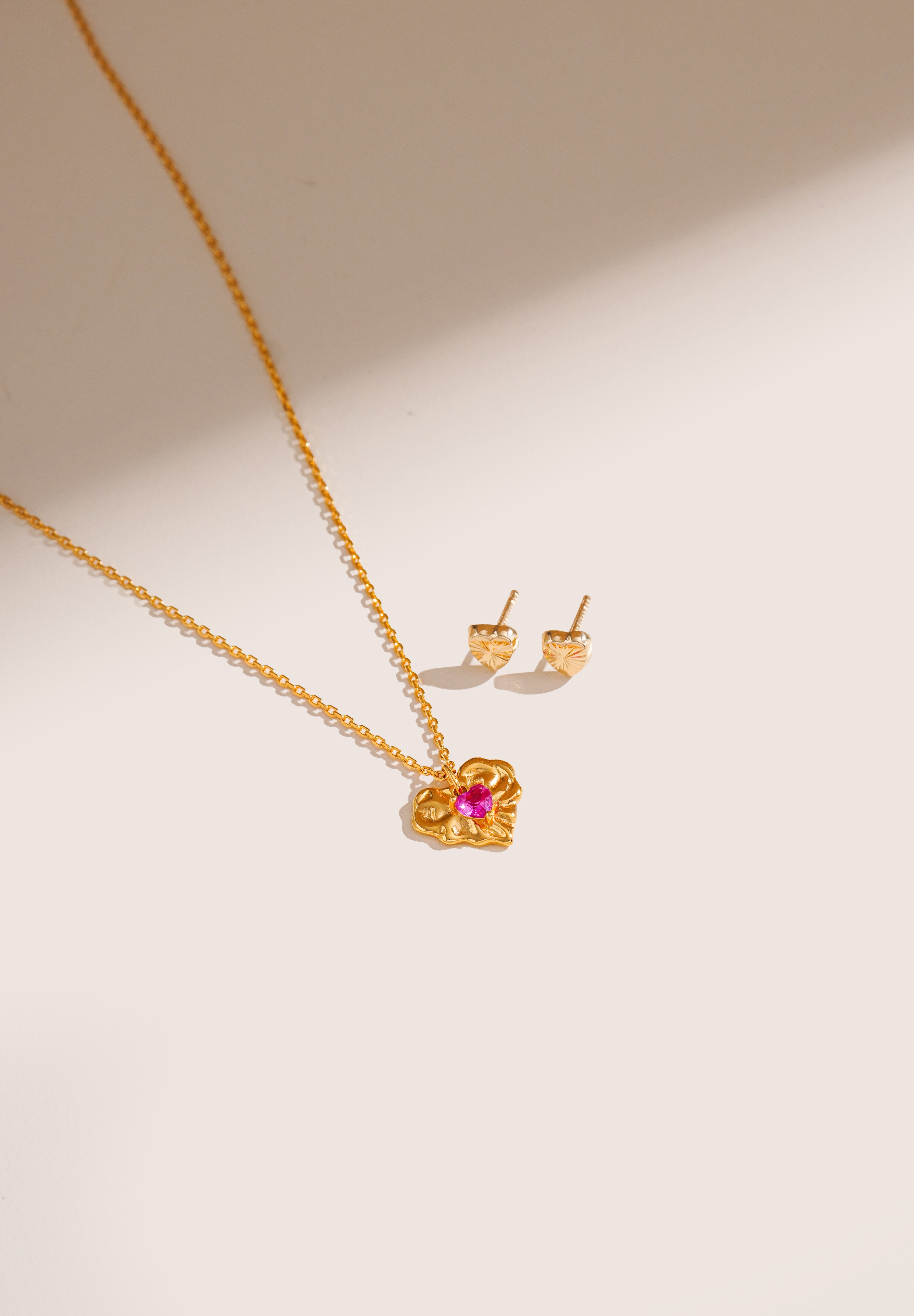Blue Francis Soho Heart Studs crafted in polished 14K gold, featuring simple heart shapes, displayed beside a gold ruby corundum necklace on a cream surface.