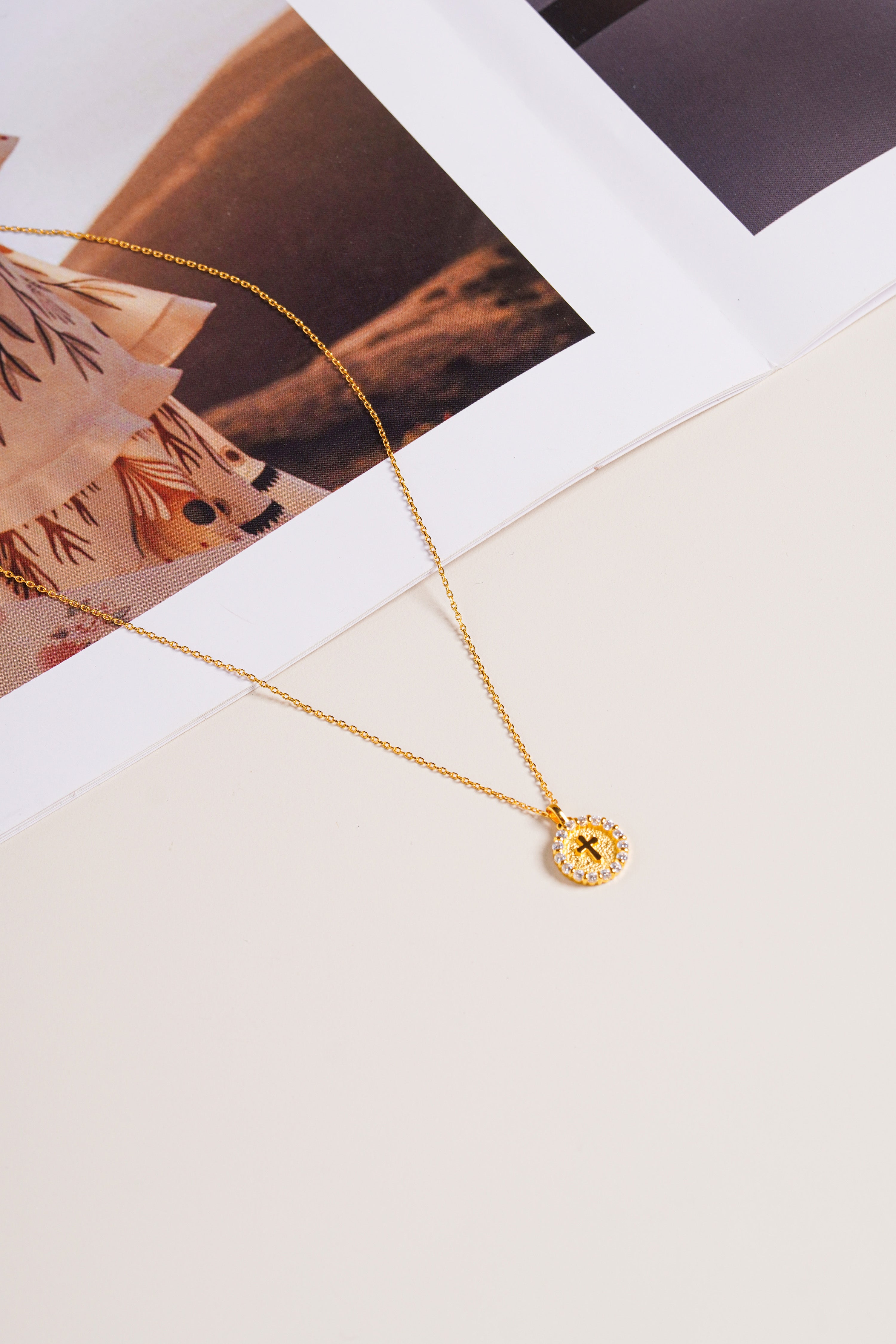 Gold vermeil Twinkle Cross Necklace with sparkling round pendant on a cream background.