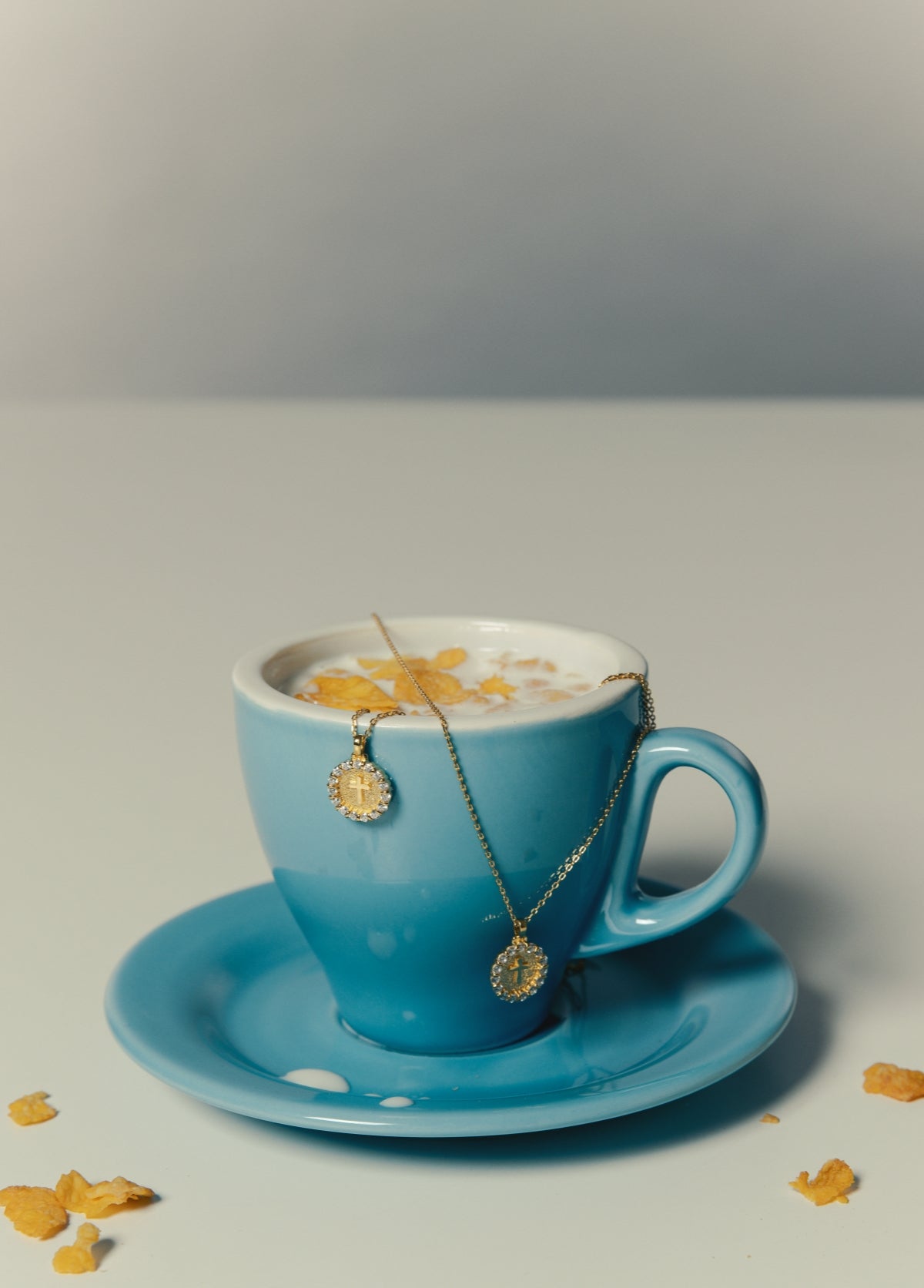 Gold vermeil necklace with round cross pendant accented by sparkling stones, draped over a blue cup of milk and cornflakes, featured for Blue Francis Shop Necklace section on the homepage.