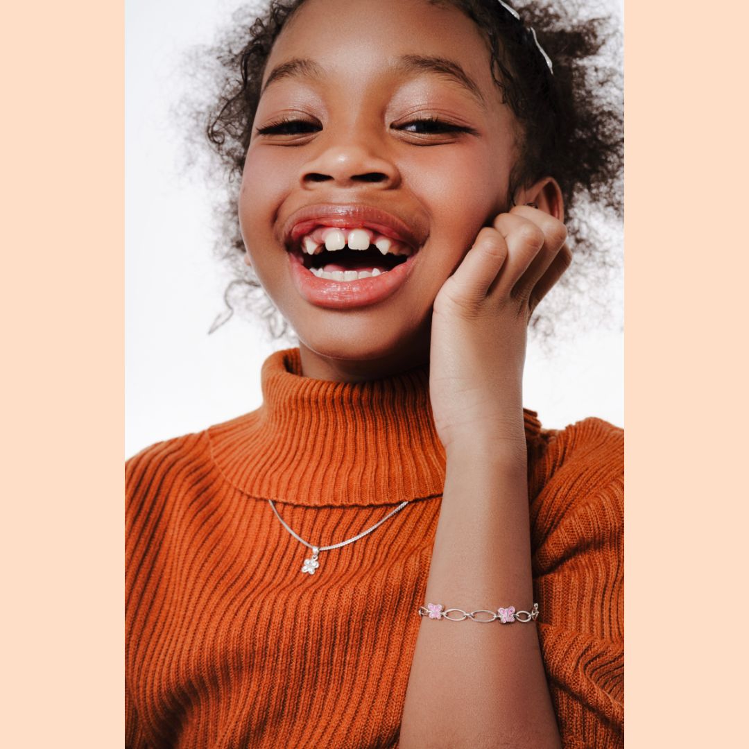 Sterling silver flower pendant necklace and butterfly bracelet from Blue Francis children's fine jewelry, styled with a rust-toned ribbed turtleneck, as featured in Earnshaw's Kids Magazine editorial, April 2026.