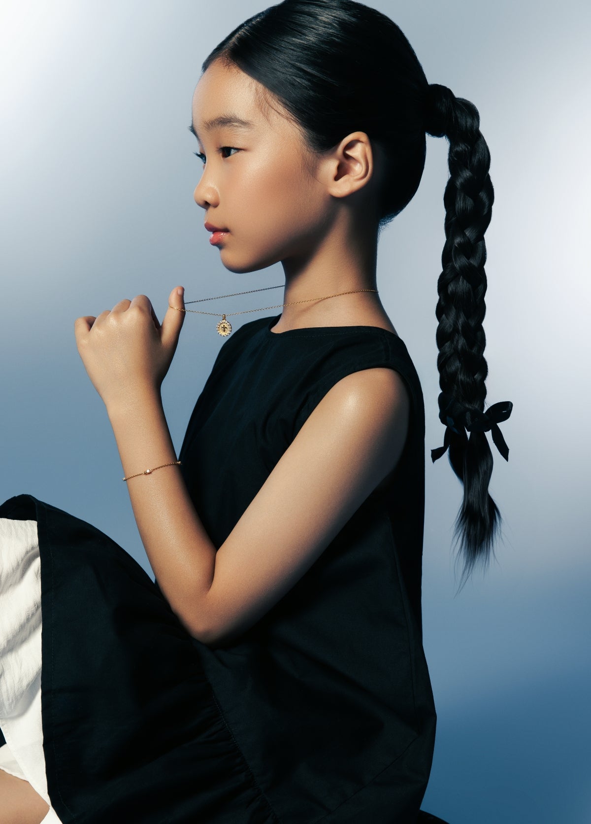 Girl in black dress holding a gold necklace pendant — Twinkle Cross Necklace and Twinkle Bracelet, Blue Francis.