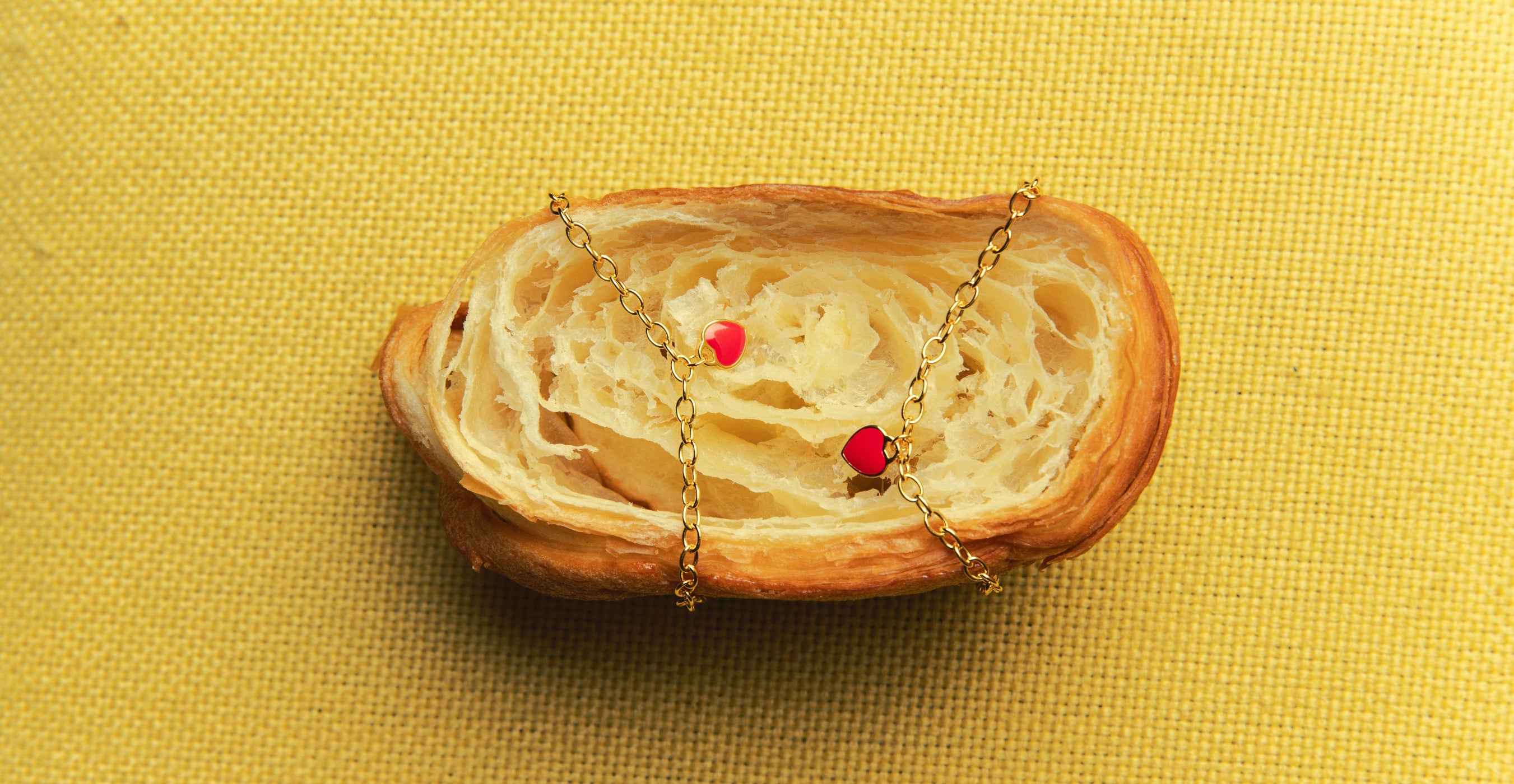 Blue Francis gold chain bracelet with red heart charms draped over a flaky croissant on a yellow textured background, used as a creative banner for the About Us page.