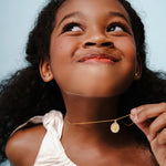 Blue Francis Kids Necklace called Cross Twinkle Necklace, made in Vermeil 18K Gold. Blue Francis model wearing a gold necklace with a pendant against a light blue background