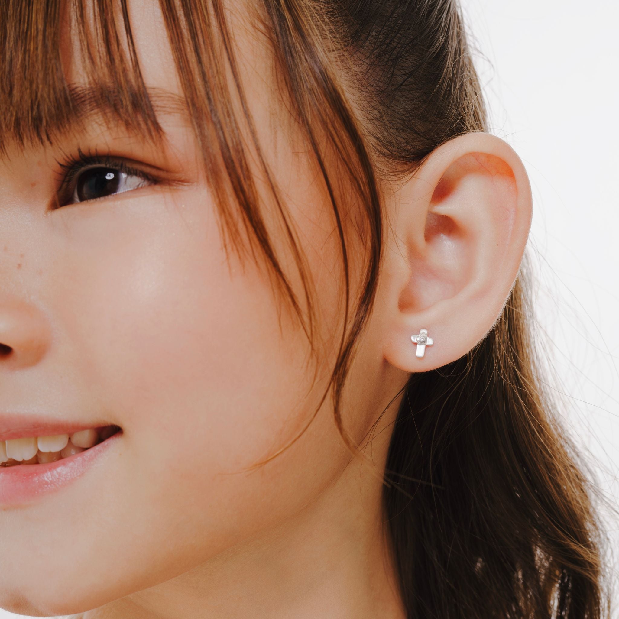 Blue Francis Kids Stud Earrings in Sterling Silver, Brooklyn Cross Earrings. A close-up picture of a person wearing a small cross silver sterling earring with a neutral background.