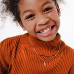 Blue Francis teens Brooklyn Cross Pendant in Sterling Silver. Blue Francis model wearing an orange sweater with a white background, wearing Brooklyn Cross Pendant and 1mm Box chain in Sterling Silver.