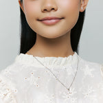 Model wears Brooklyn Cross Necklace with small heart center and sterling silver cubic zirconia studs, smiling gently on neutral background for detail.