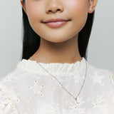 Model wears Brooklyn Cross Necklace with small heart center and sterling silver cubic zirconia studs, smiling gently on neutral background for detail.