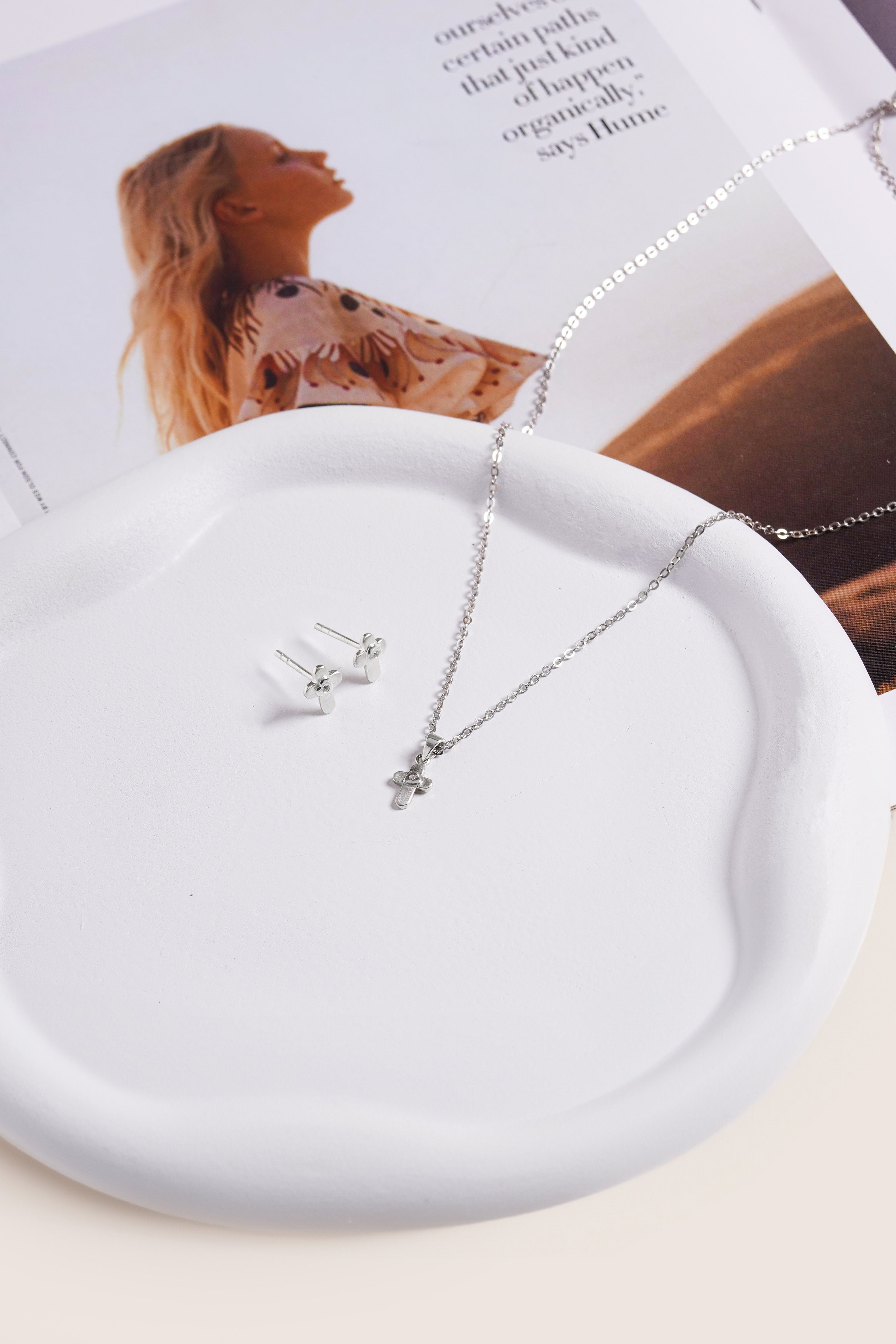 Brooklyn Cross Necklace in sterling silver shown draped on a white ceramic dish next to cross stud earrings, styled with a fashion magazine.