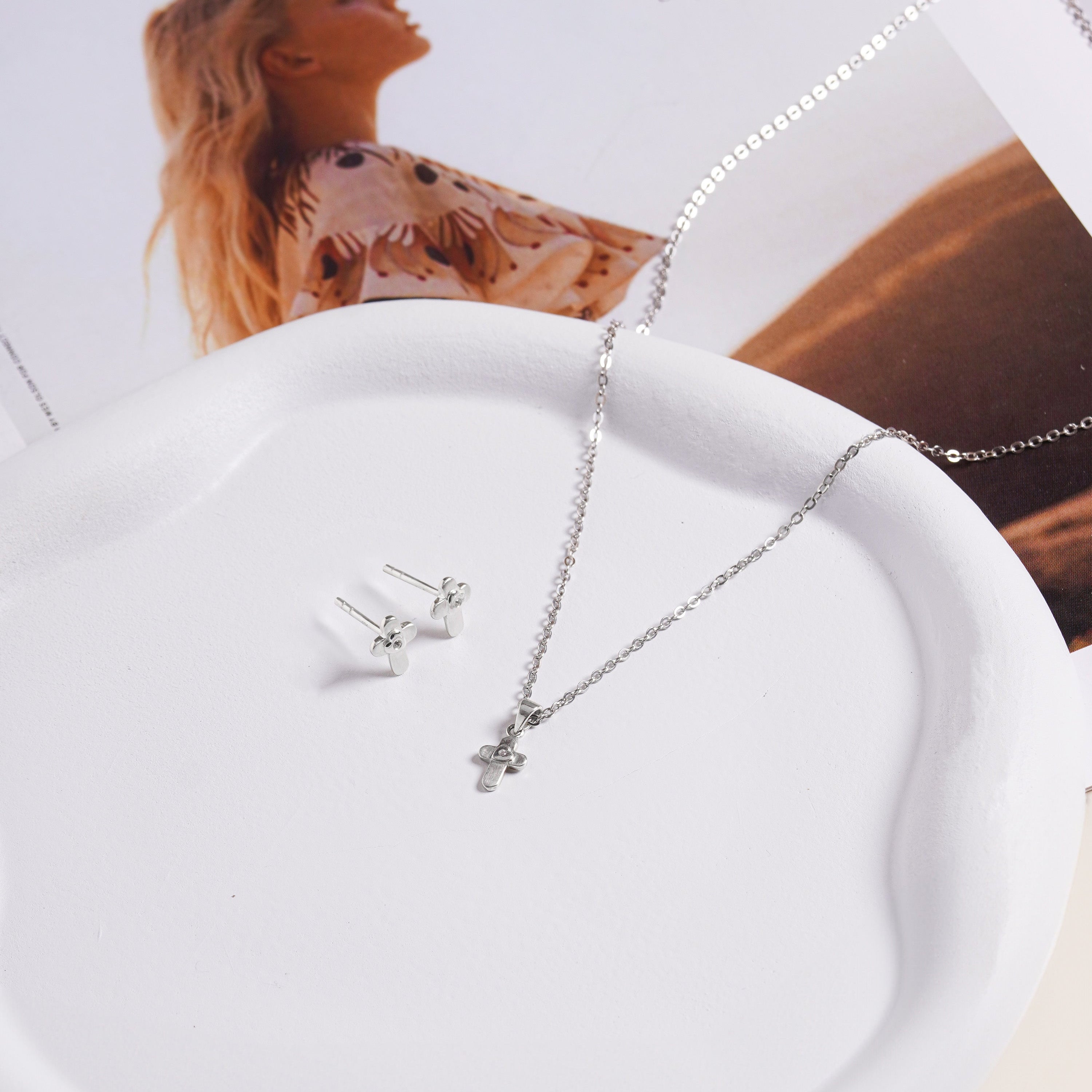Brooklyn Cross Studs in sterling silver displayed on a white ceramic dish beside a silver cross necklace, styled with a magazine.