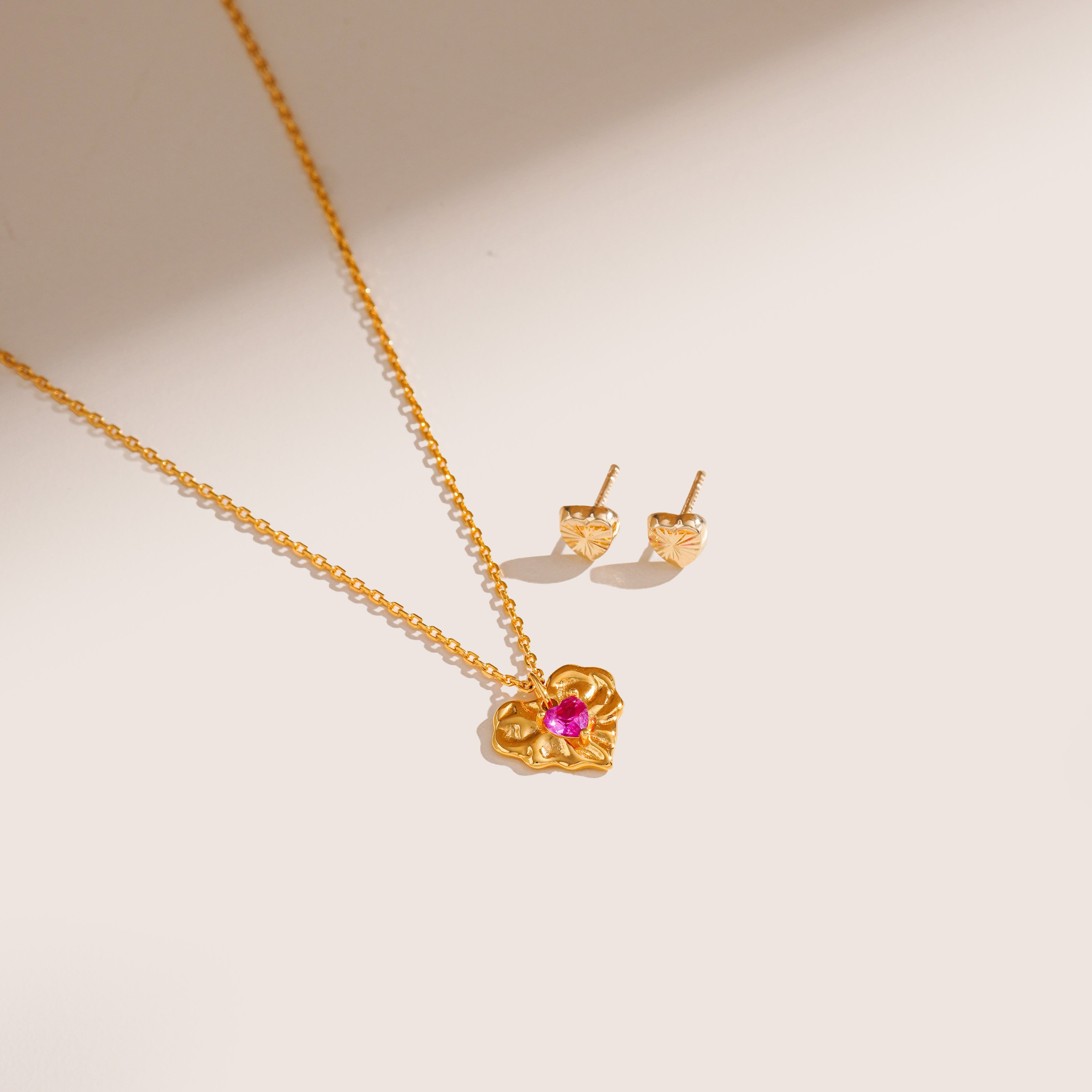 Blue Francis Soho Heart Studs crafted in polished 14K gold, featuring simple heart shapes, displayed beside a gold ruby corundum necklace on a cream surface.