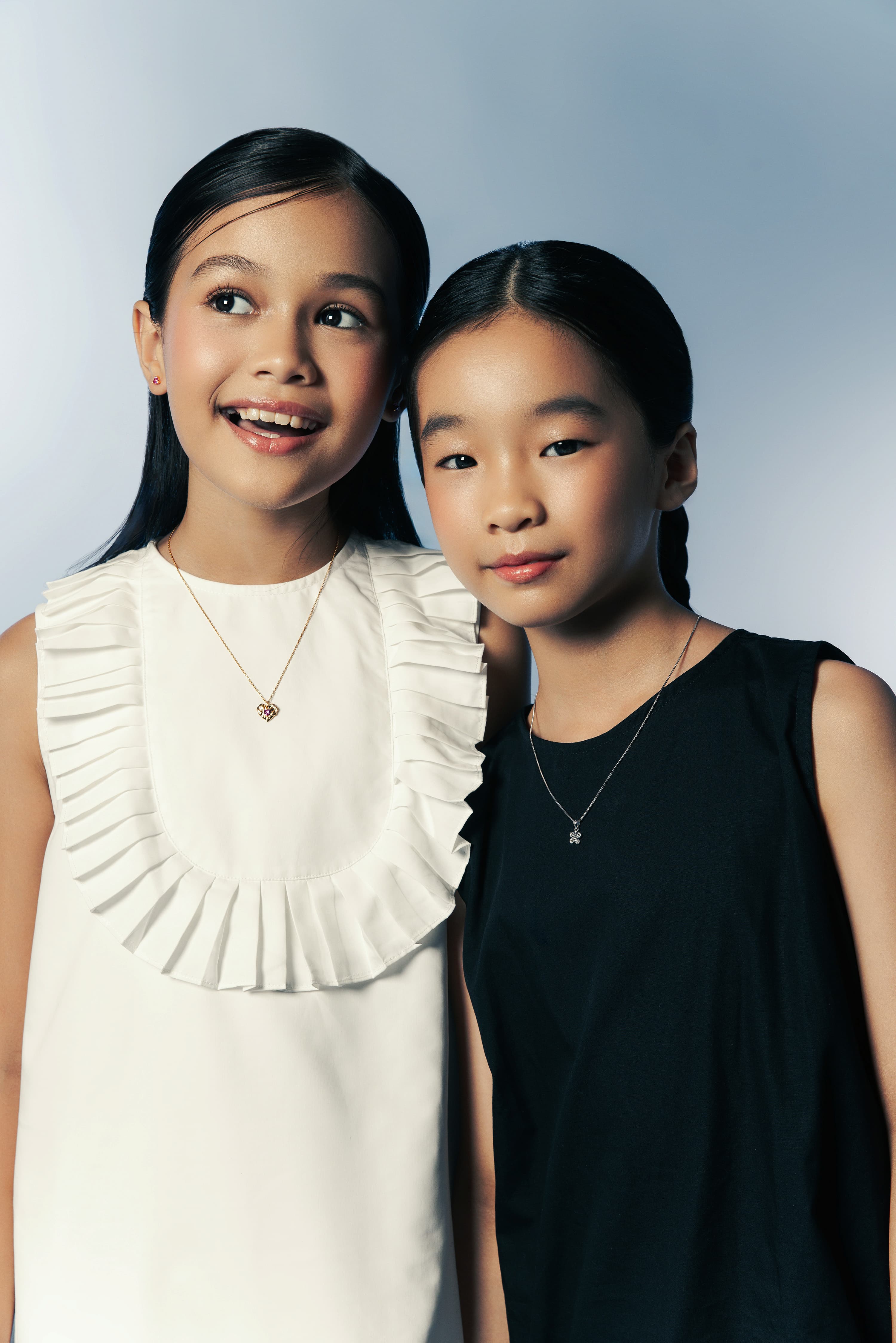 Two young girls wearing Blue Francis jewelry, one in a white dress with a gold heart necklace and red stud earrings, and one in a black dress with a silver cross necklace, against a soft blue gradient background.