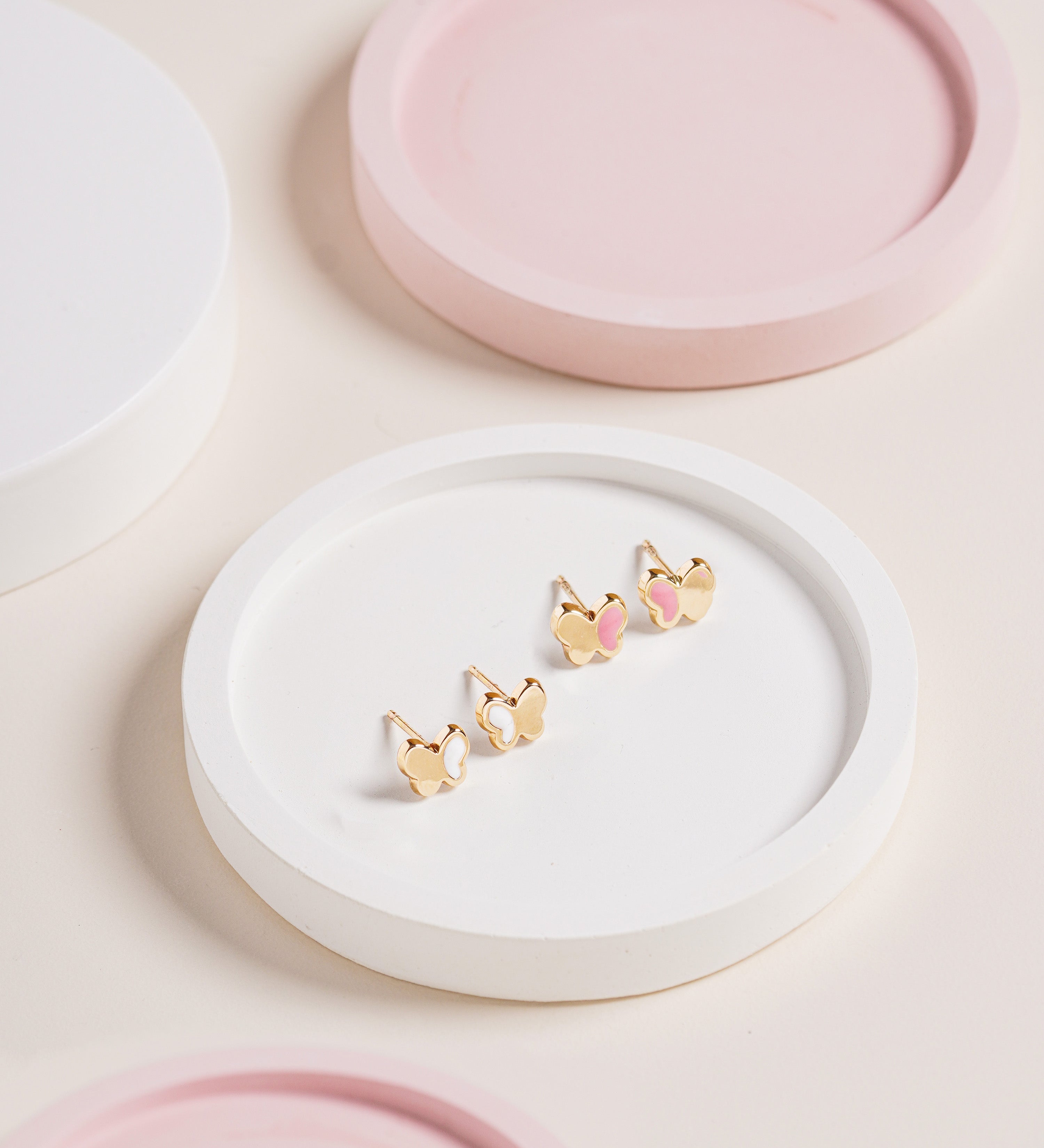 14K gold butterfly stud earrings with pink and white enamel accents, displayed on a white tray with soft pastel pink decor, featured in Blue Francis Homepage Shop Earrings section.