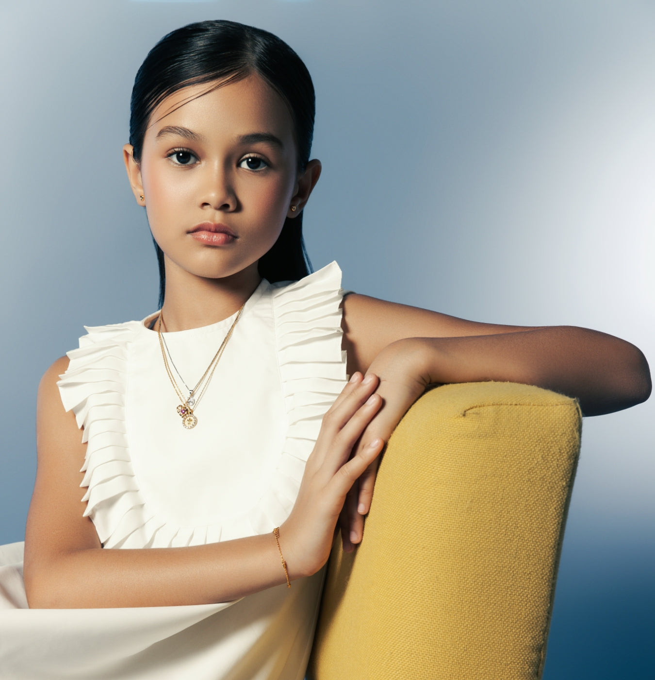 A young girl wearing layered gold necklaces and a bracelet from Blue Francis, seated on a yellow chair in soft light.