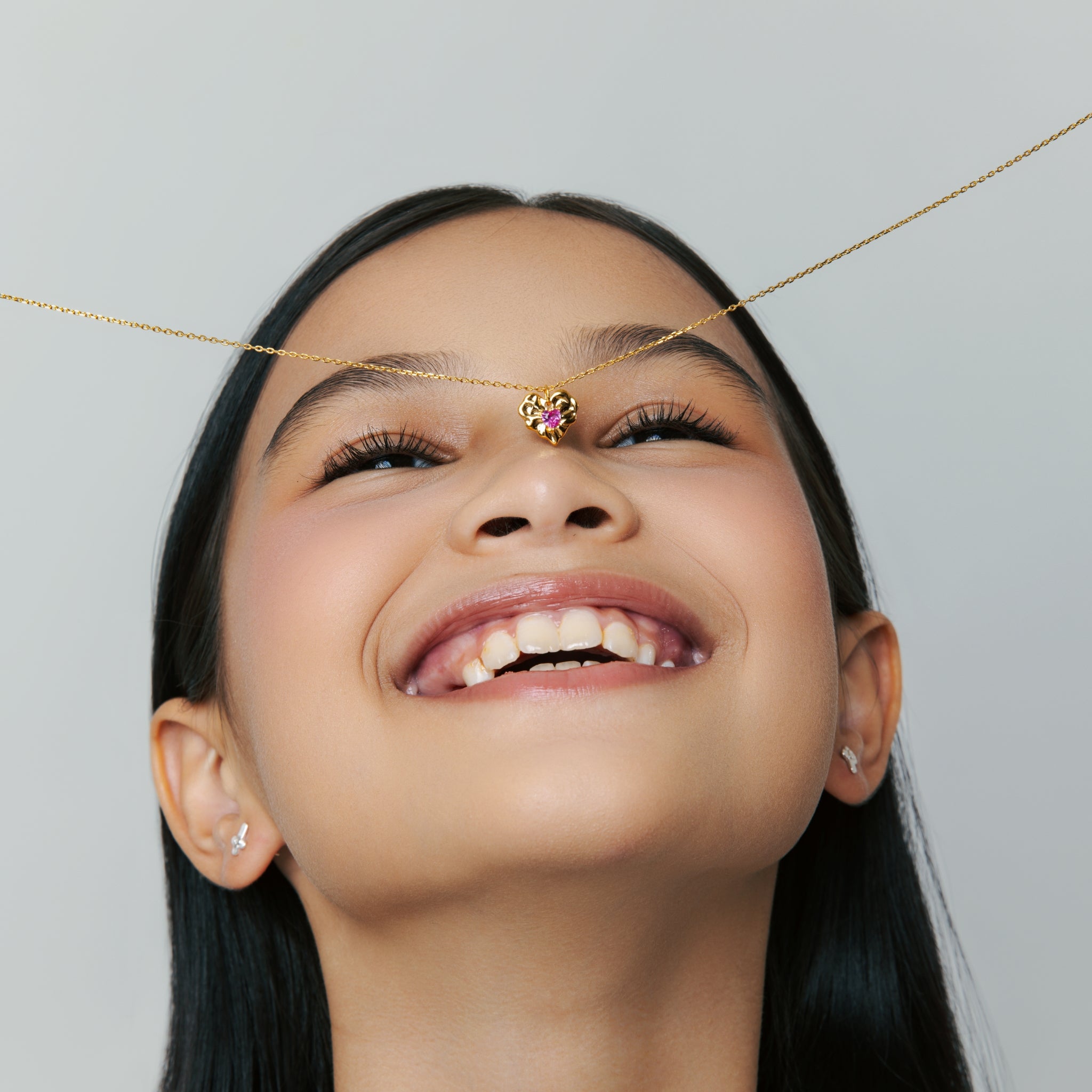 Blue Francis Ruby Liberty Necklace in gold vermeil with a heart-shaped pendant set with a ruby corundum stone, shown playfully held across a smiling model's face.